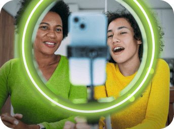 Two people sit in front of a green o-ring light, smiling & speaking into a phone camera.